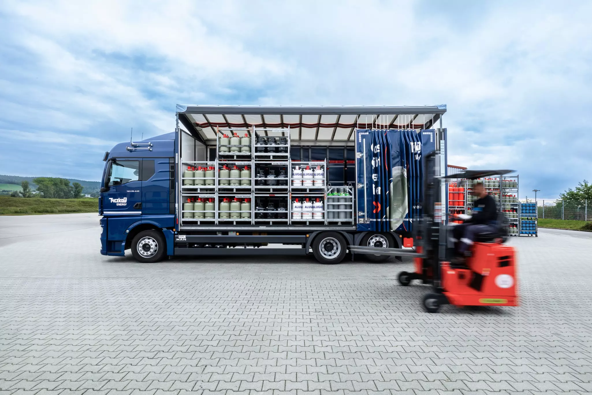 Flaschengas Transport - seitliche Ansicht eines Flaschengas LKWs mit offener Plane und mehreren Paletten mit verschiedenen Gasflaschen auf der Ladefläche, davor ein fahrender Gabelstapler